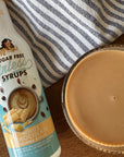 A bottle of Mary's Sugar Free Sinless Syrups White Chocolate Coffee Syrup next to a glass filled with the light brown coffee syrup, set on a wooden table with a striped cloth background.