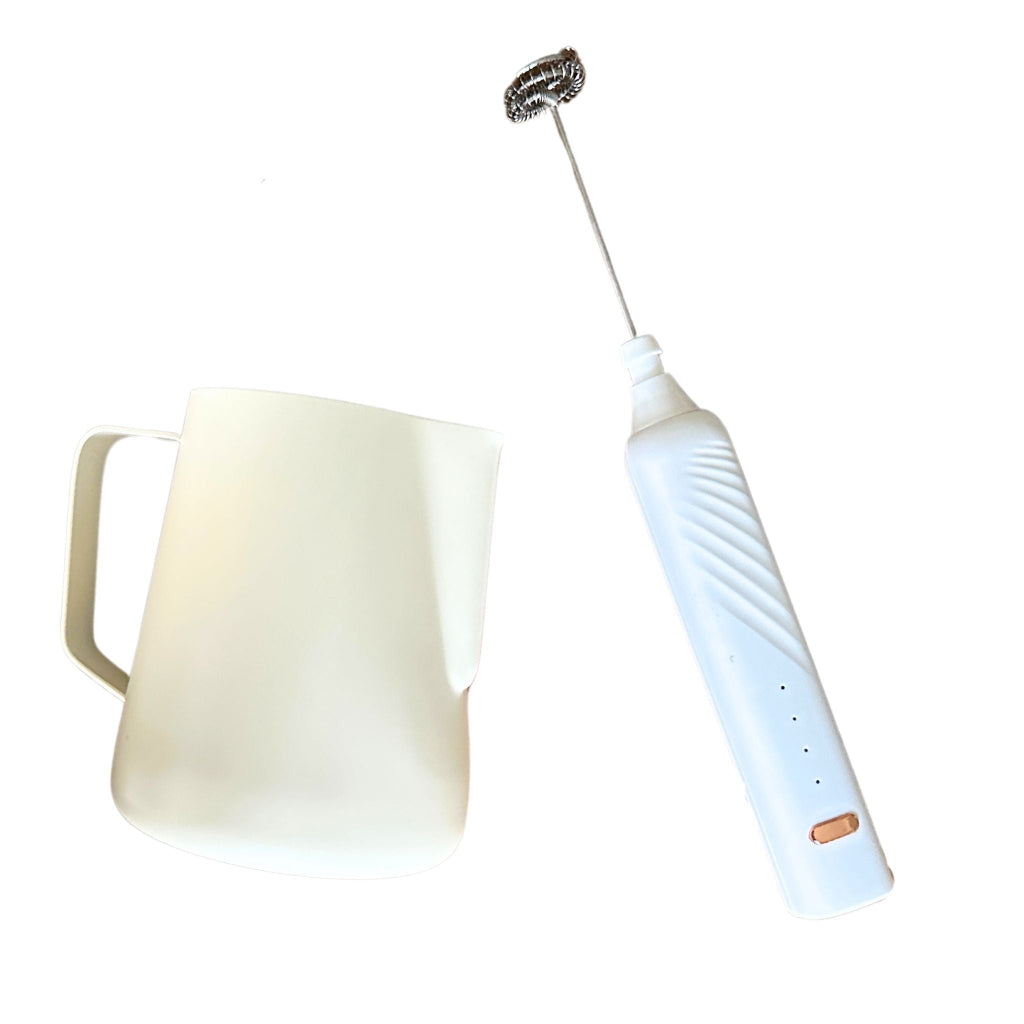 A stainless steel frothing pitcher next to an electric frother wand with a white handle and a USB charger.